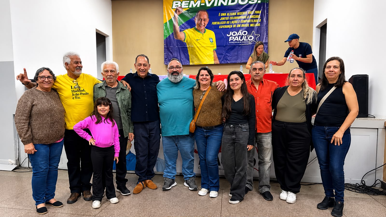 João Paulo Cunha posa ao lado de participantes de um encontro, com uma criança e um painel de boas-vindas ao fundo.
