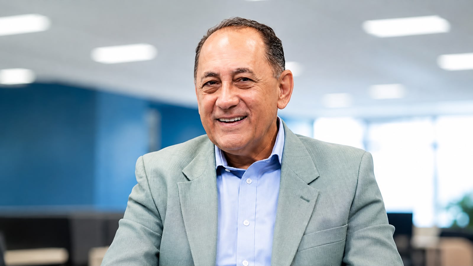 João Paulo Cunha sorrindo, sentado em um ambiente interno, usando terno claro e camisa azul.