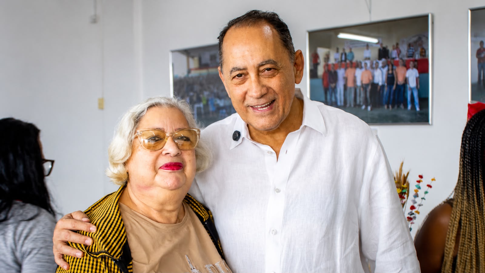 João Paulo Cunha aparece sorrindo, usando camisa branca, ao lado de uma apoiadora durante um encontro. Ao fundo, há fotografias de atividades coletivas e outros participantes do evento.