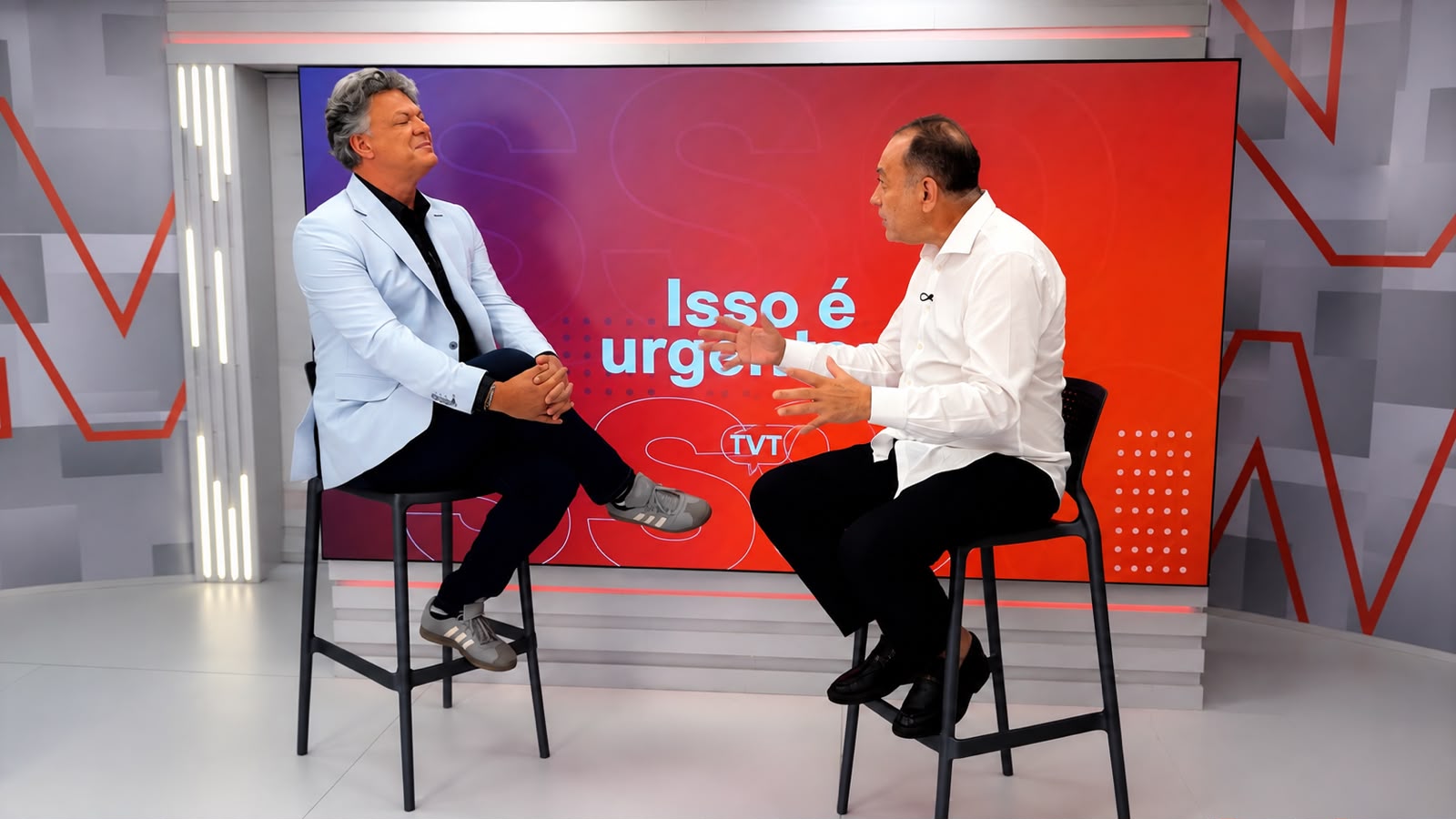 João Paulo Cunha, de camisa branca, conversa com o apresentador Don Ernesto durante entrevista no programa Isso é Urgente, da TVT News. Os dois estão sentados em bancos no estúdio, diante de um painel vermelho com o nome do programa.
