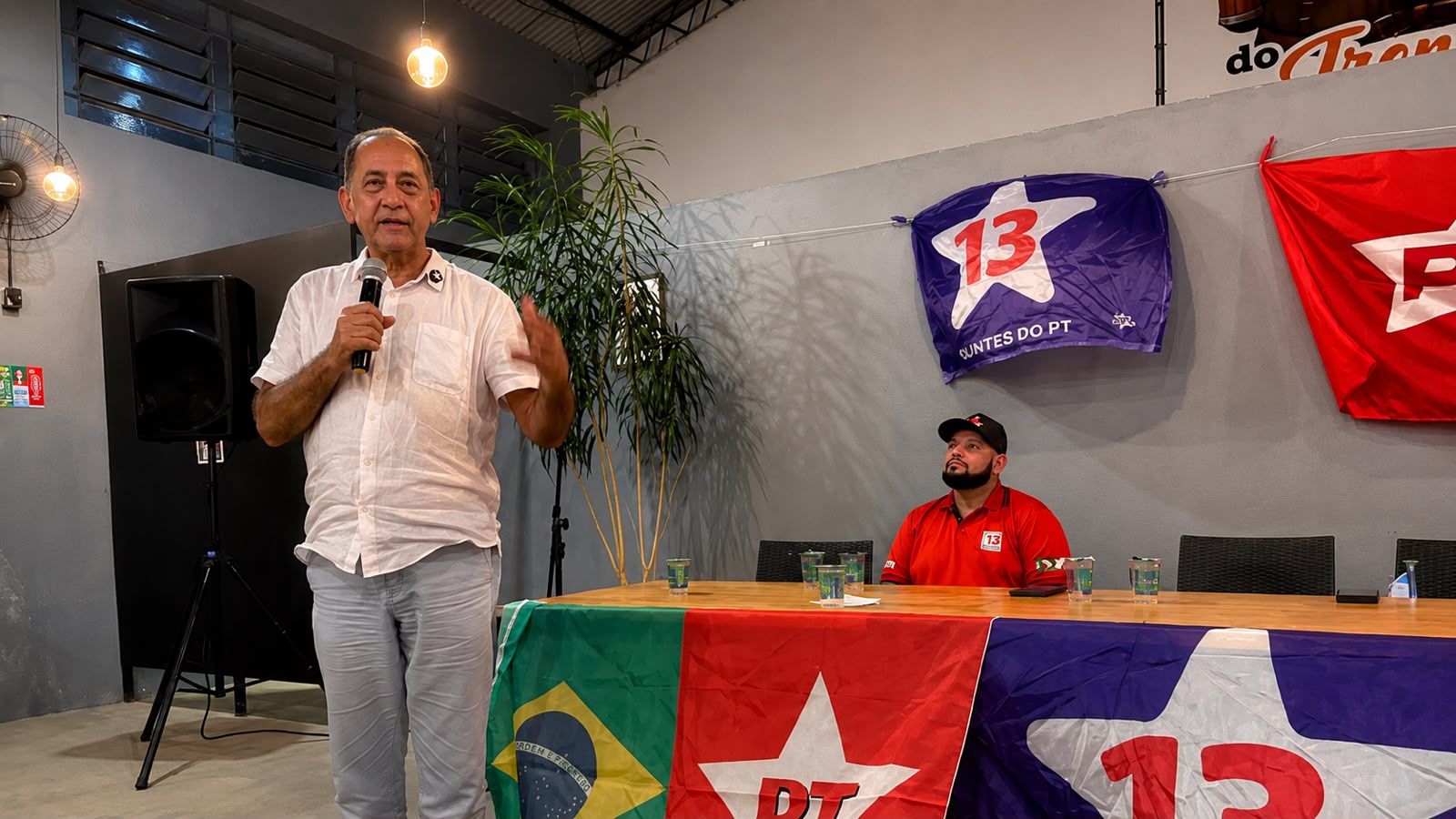 Homem de camisa branca fala ao microfone durante encontro do PT, enquanto outro homem acompanha sentado à mesa, cercado por bandeiras do partido.