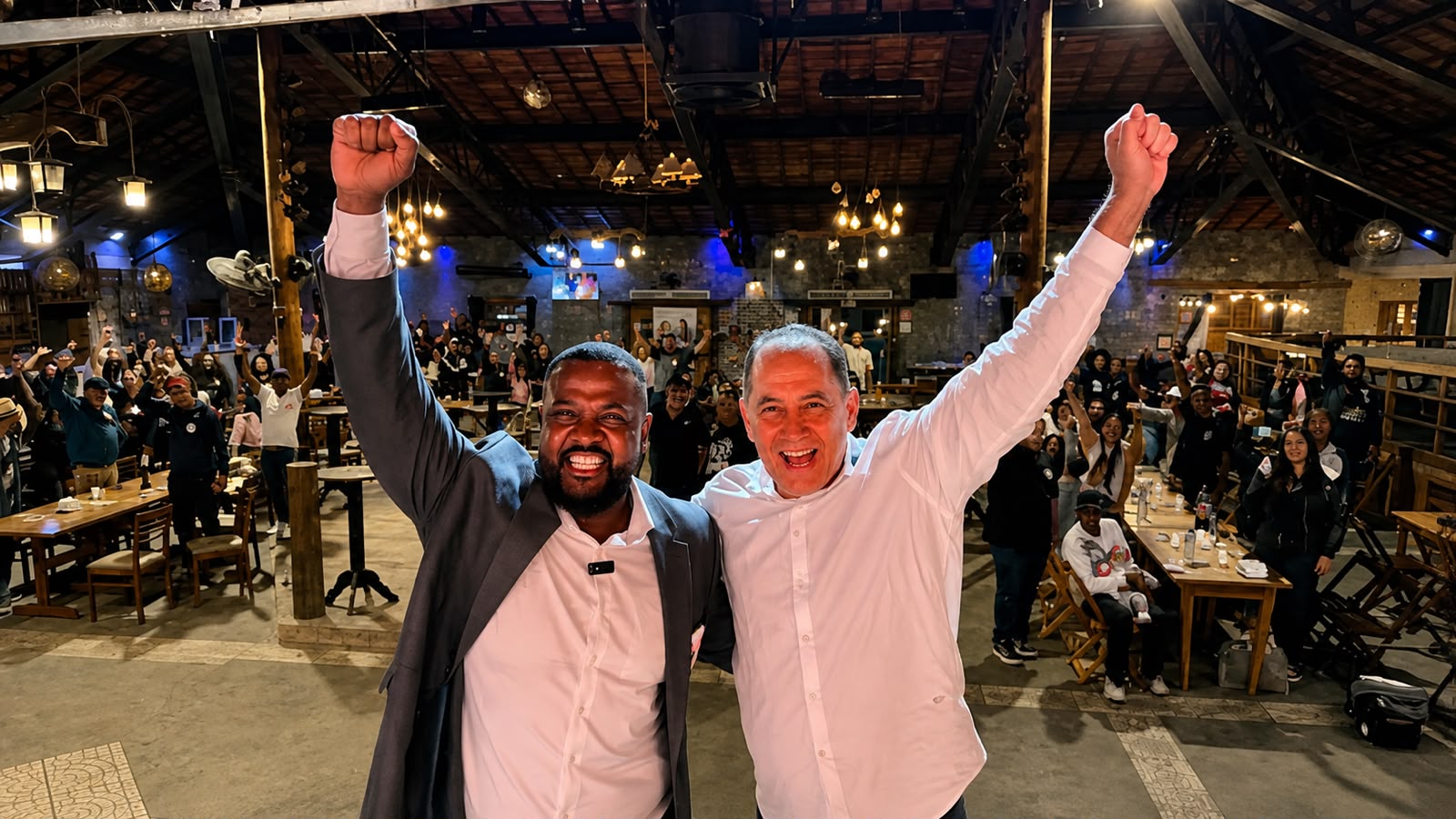João Paulo aparece ao lado de uma liderança durante encontro com apoiadores. Os dois estão sorrindo e com os braços levantados, enquanto o público ao fundo acompanha o momento em um espaço de confraternização.