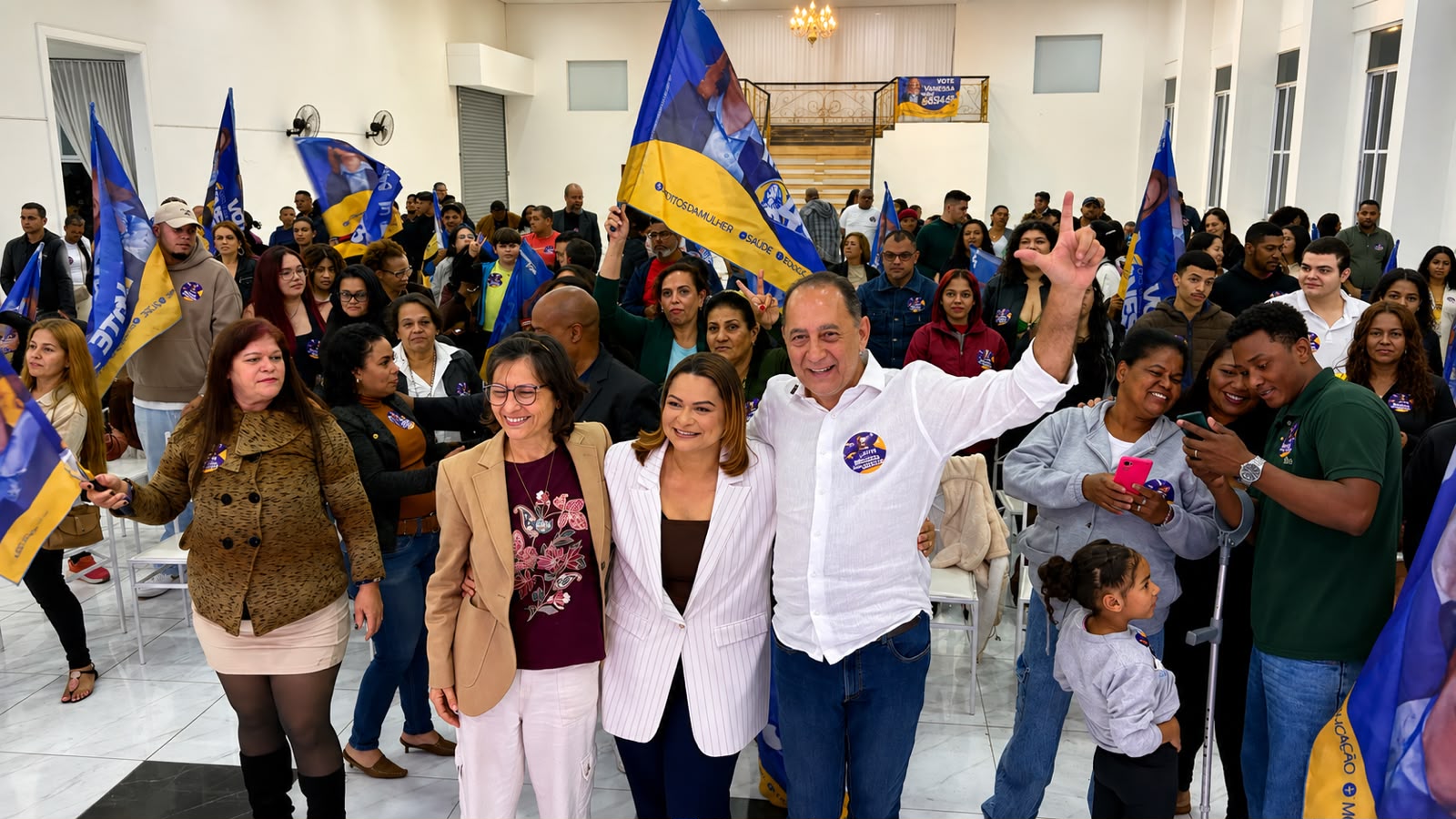 João Paulo e Vanessa Maia aparecem lado a lado em evento político lotado, cercados por apoiadores que seguram bandeiras da campanha. João Paulo levanta o braço enquanto o público acompanha o encontro.