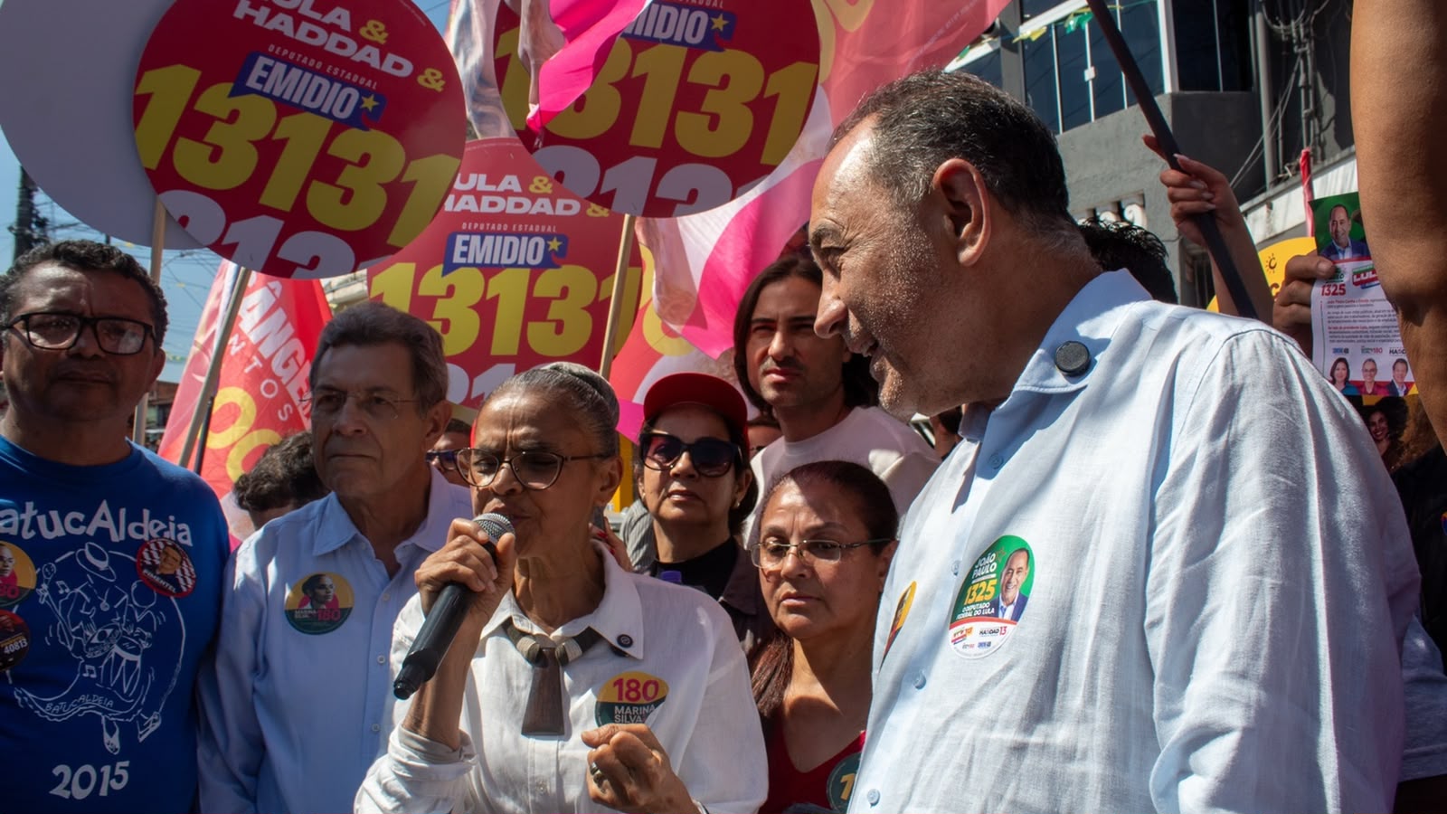 João Paulo e Marina Silva aparecem durante caminhada política em meio a apoiadores. Marina segura um microfone e fala ao público, enquanto João Paulo a acompanha ao lado de outras lideranças e bandeiras de campanha.