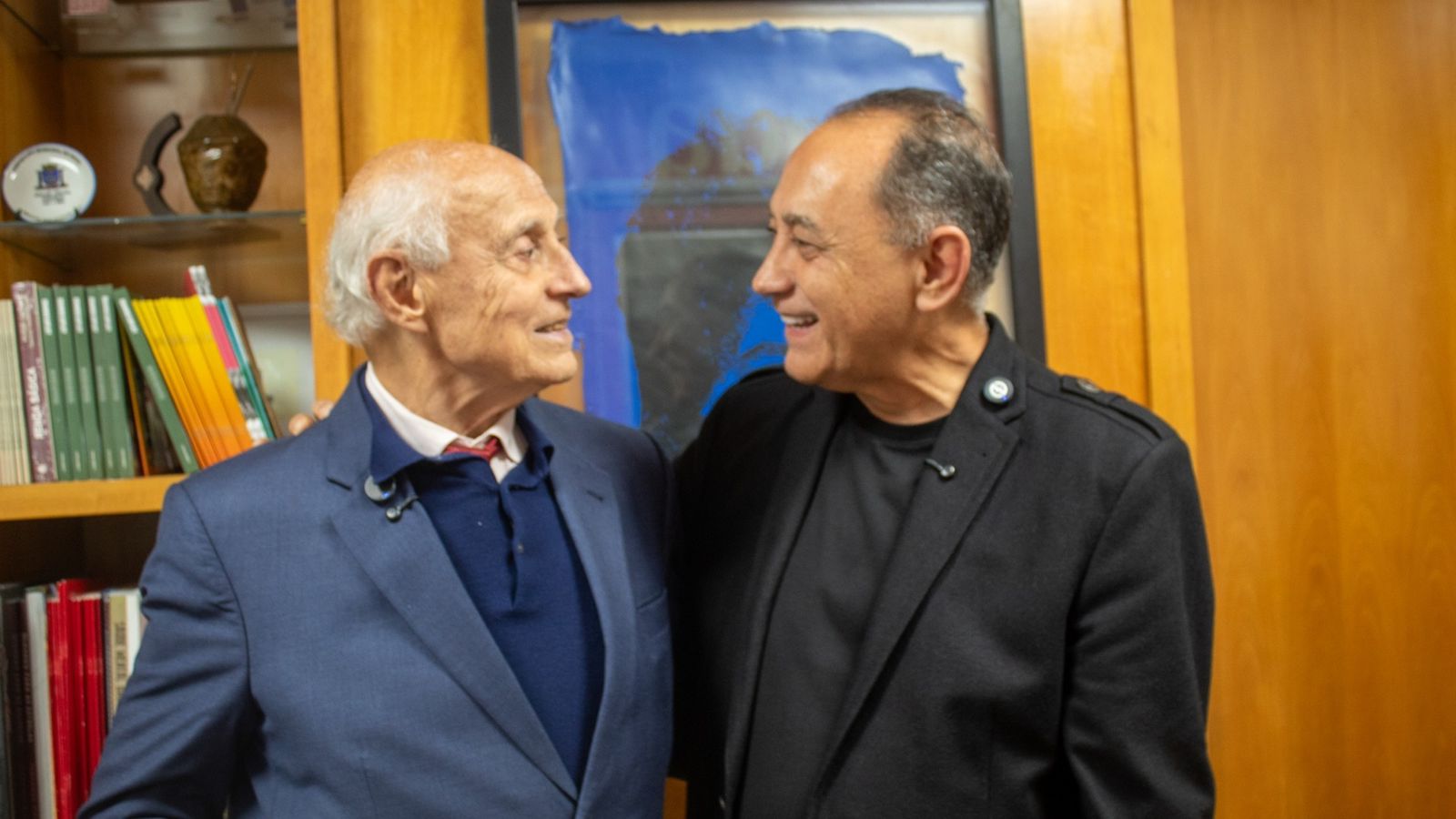 Dois homens conversam e posam juntos diante de uma estante de livros.