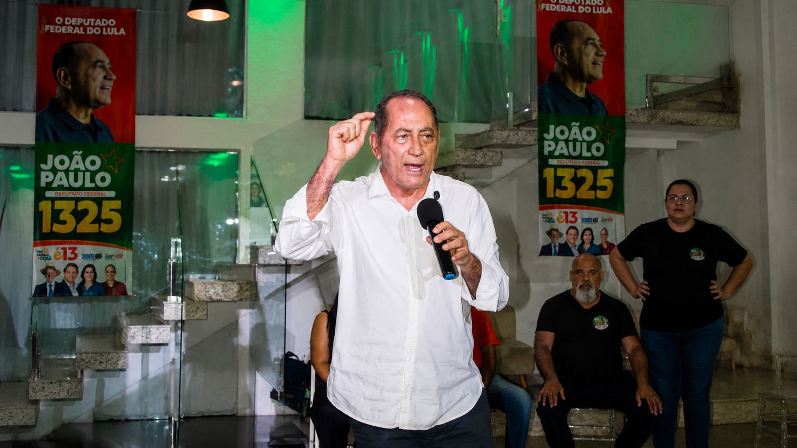 João Paulo fala ao microfone durante atividade em Mutinga, diante de moradores e apoiadores, com cartazes de sua candidatura ao fundo.