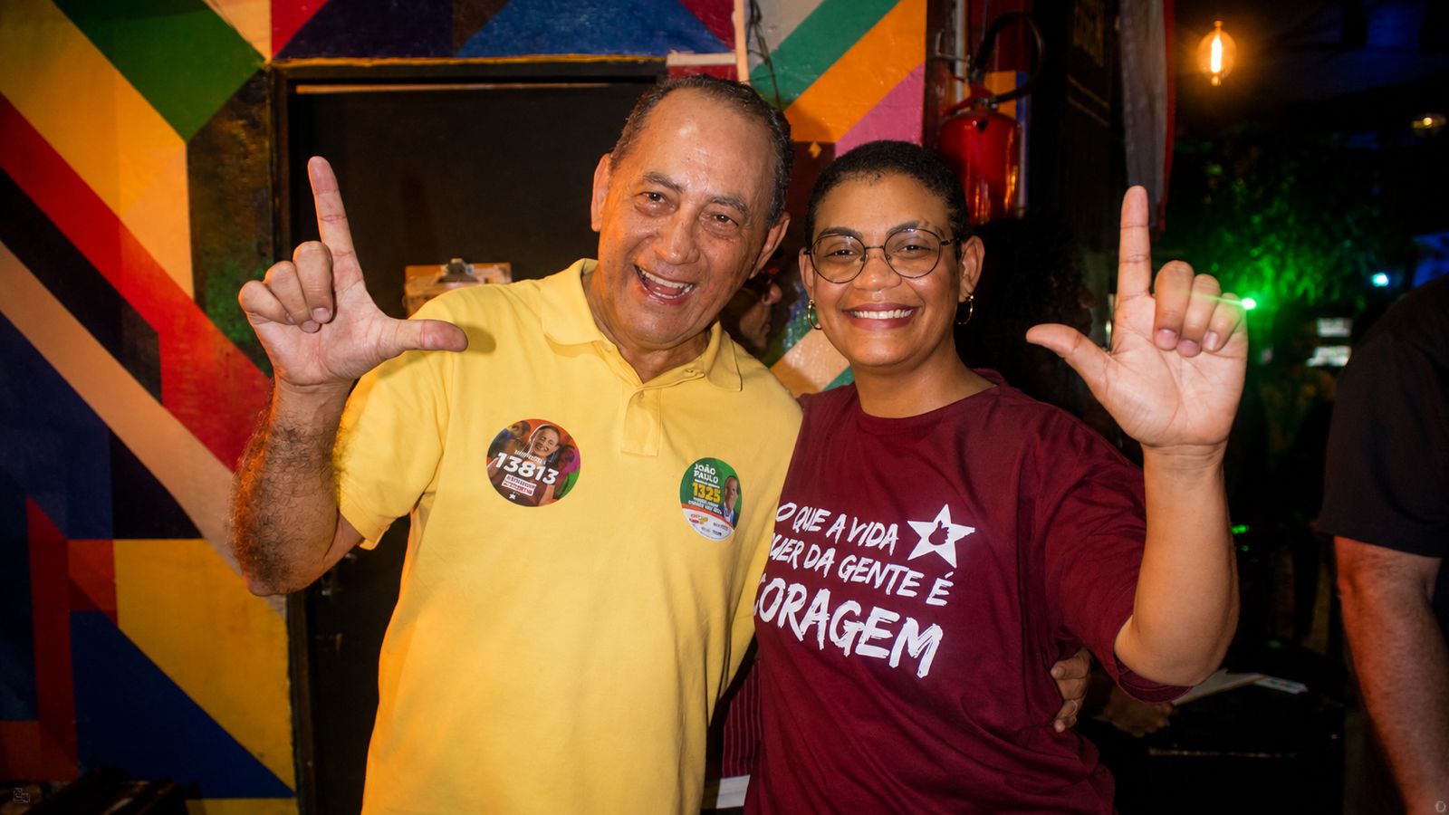 João Paulo participa de encontro com apoiadores em Guarulhos João Paulo aparece ao lado de uma mulher, ambos sorrindo e fazendo um gesto com as mãos para a câmera. Ao fundo, há uma parede com pintura colorida e outras pessoas no ambiente. Os dois usam adesivos relacionados à campanha eleitoral.