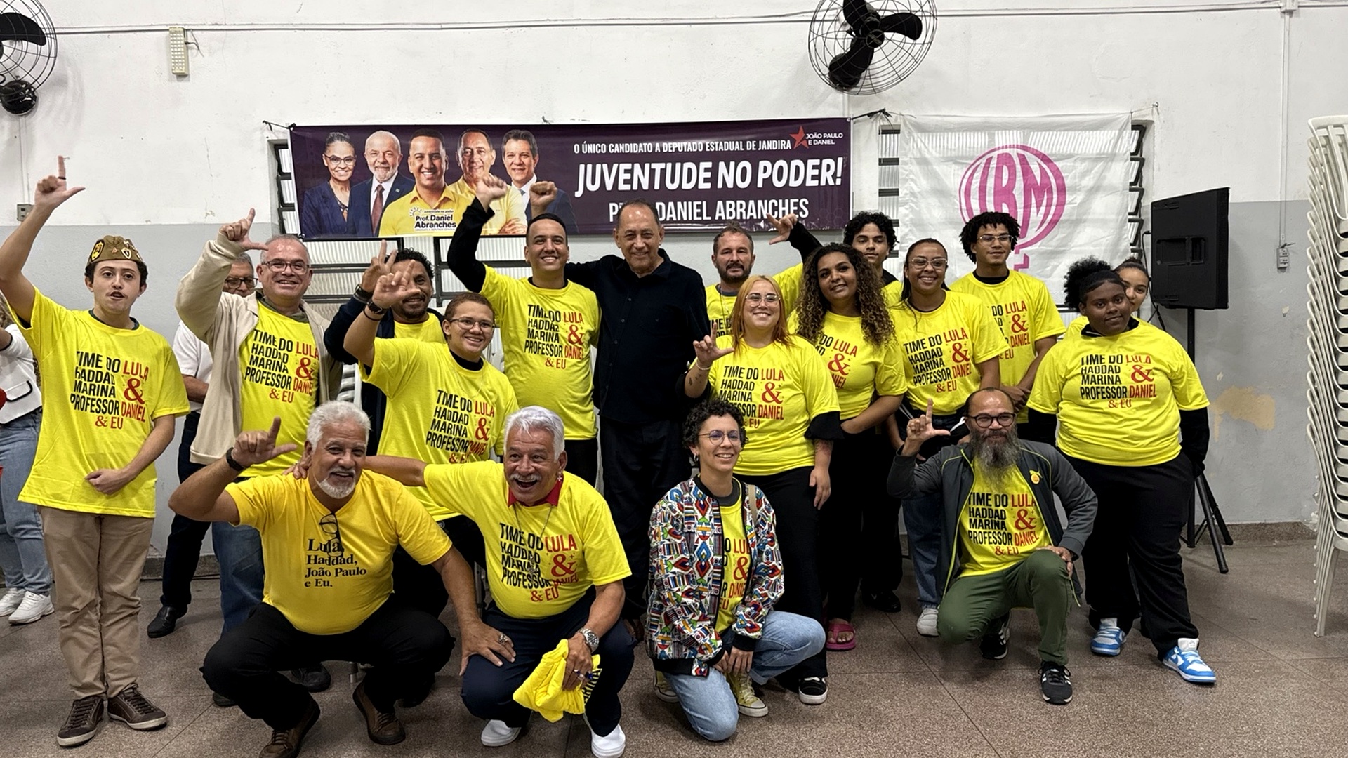 João Paulo Cunha posa ao lado de participantes durante encontro político, com pessoas usando camisetas amarelas e uma faixa de campanha ao fundo