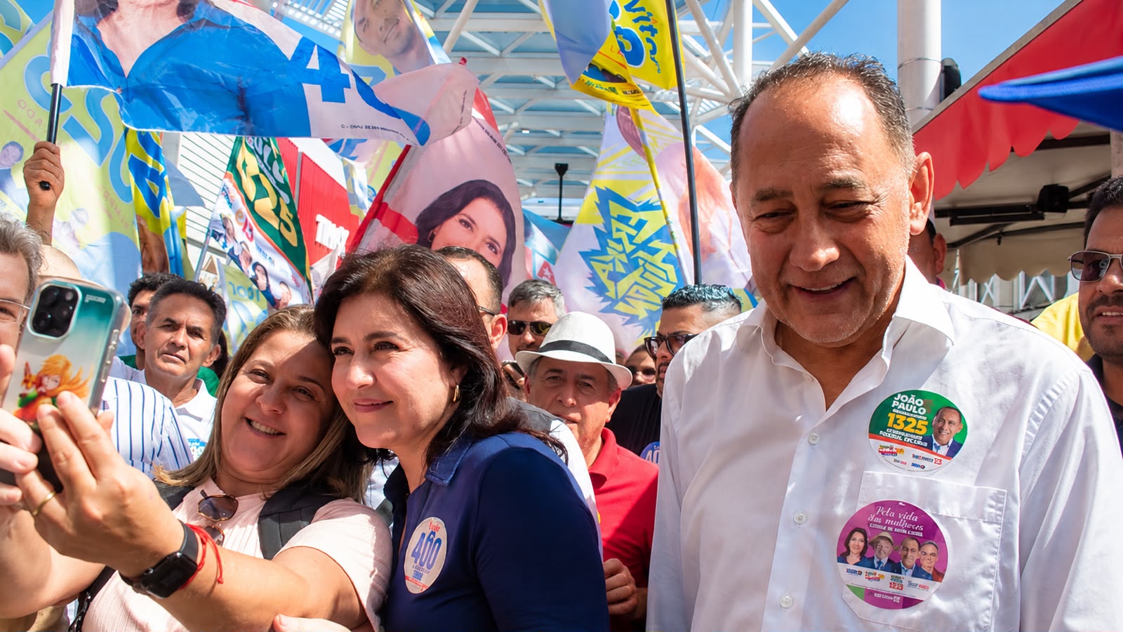 Simone Tebet aparece ao lado de João Paulo Cunha durante caminhada em Osasco, enquanto uma apoiadora registra o momento com o celular.