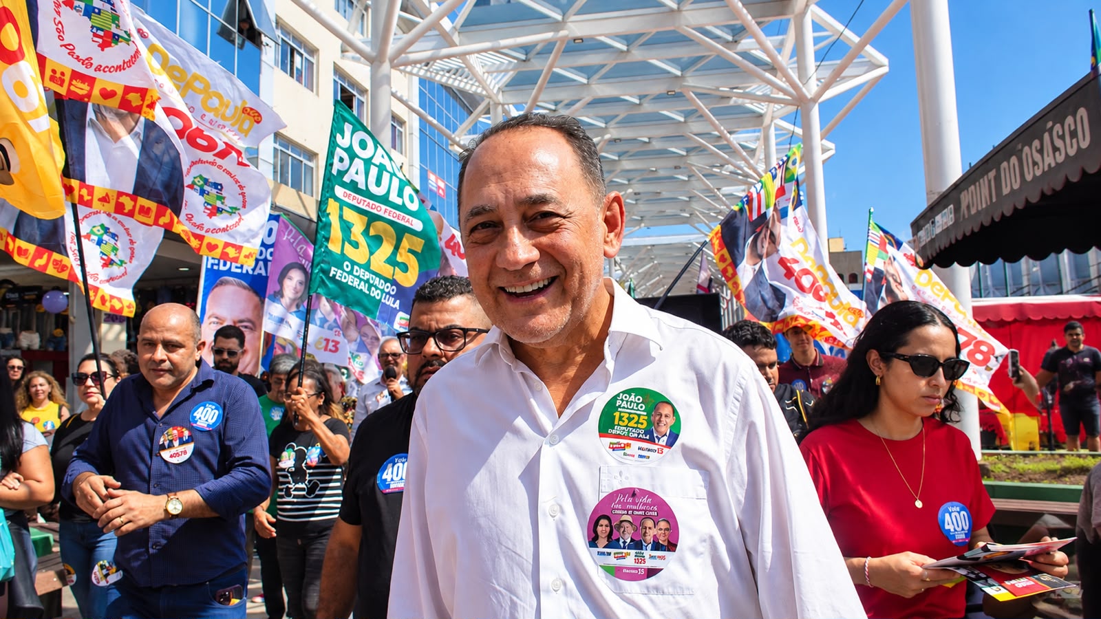 João Paulo Cunha aparece em destaque durante caminhada política em Osasco, cercado por apoiadores e bandeiras de campanha.