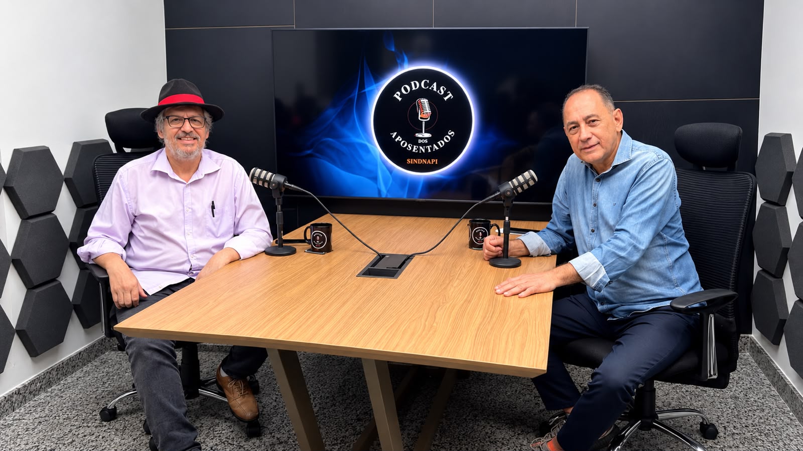 João Paulo Cunha participa do Podcast dos Aposentados Duas pessoas participam de uma gravação no estúdio do Podcast dos Aposentados, sentadas diante de microfones e de uma mesa de madeira.